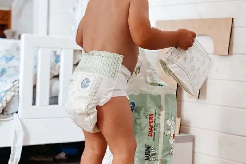 Un bimbo con un pannolino indosso, vicino a una confezione di pannolini. Sembra che i pannolini scadano, ma non c'è fretta.