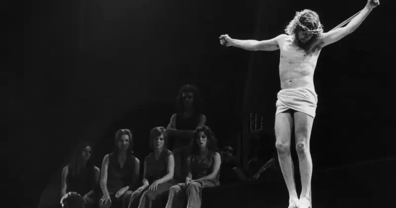 Scena z musicalu Jesus Christ Superstar: Jezus na krzyżu, otoczony przez postaci, w tym kobiety siedzące wpatrzone w niego.