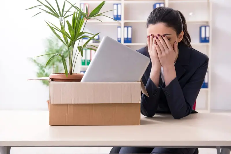 Verzweifelte Frau mit Karton und Laptop im Büro. Sie muss eine innerbetriebliche Versetzung ablehnen und ist sichtlich mitgenommen.