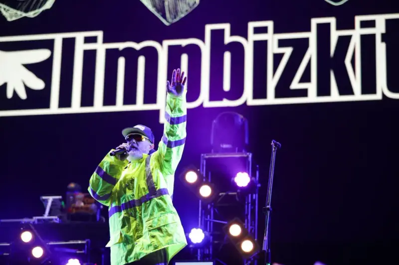 Fred Durst z Limp Bizkit na koncercie, w neonowej kurtce, z ręką w górze. Tło to logo zespołu.