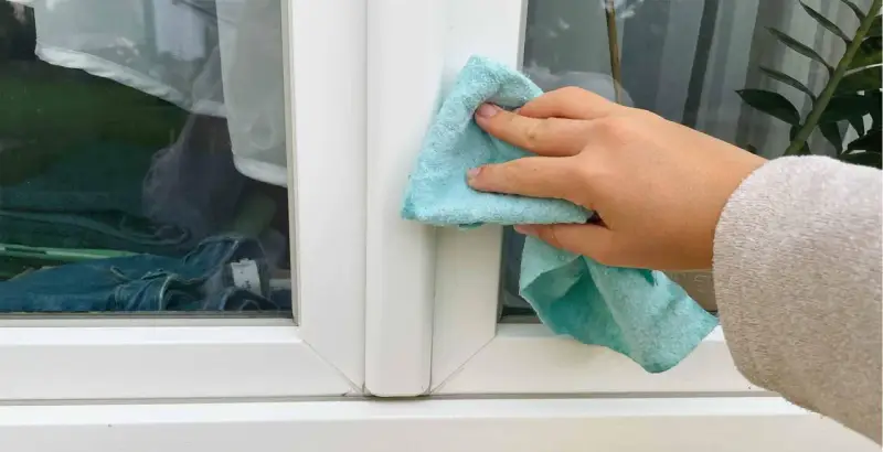 Hand reinigt mit blauem Tuch den weißen Rahmen eines Kunststofffensters.