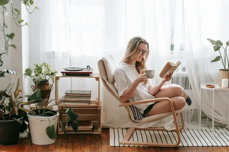 Mujer leyendo en un sillón, taza en mano, rodeada de plantas y libros. Un rincón de lectura con decoración acogedora y un tocadiscos.