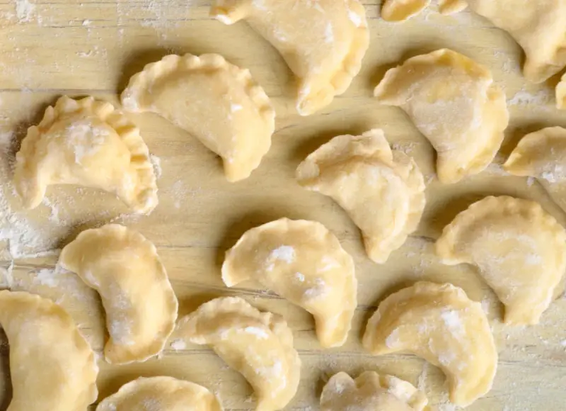 Surowe pierogi z farszem na pierogi ruskie, ułożone na drewnianym blacie, posypane mąką.