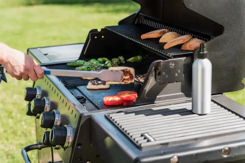 Ręka z szczypcami obraca na grillu kawałki sera halloumi, obok pieczone pomidory i papryczki.