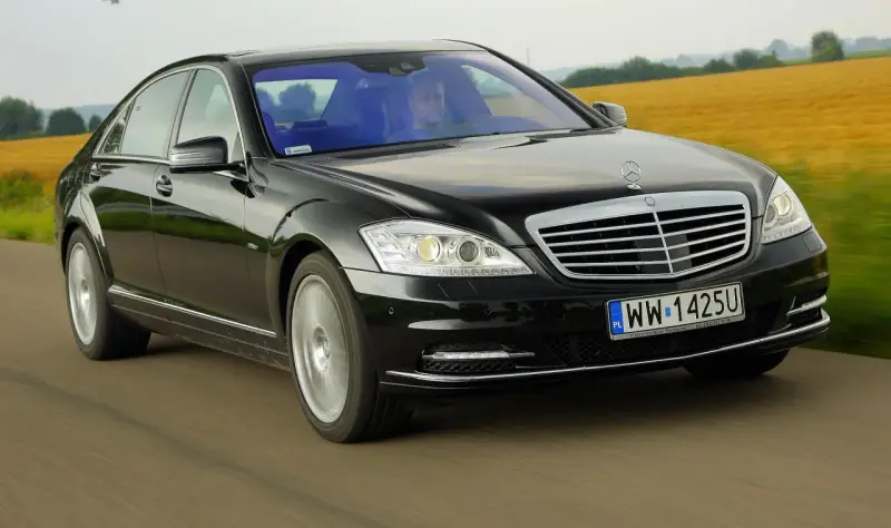 Czarny Mercedes-Benz W221 pędzi po drodze, mijając pola.