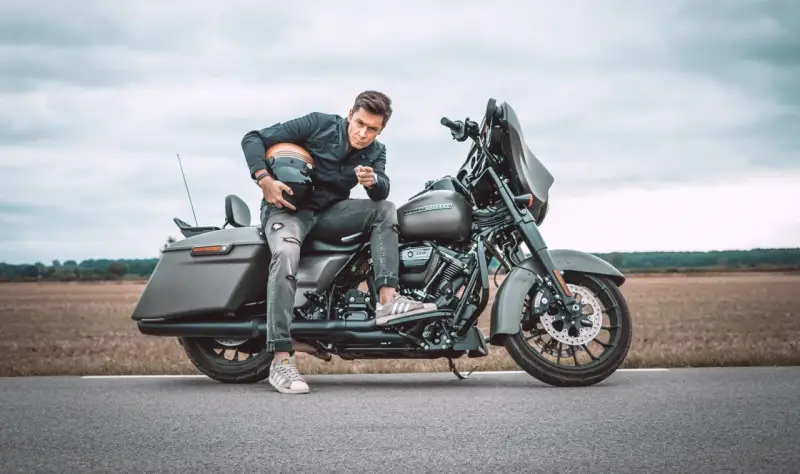 Mężczyzna w czarnej kurtce i podartych jeansach siedzi na motocyklu Harley-Davidson Street Glide, trzymając kask.