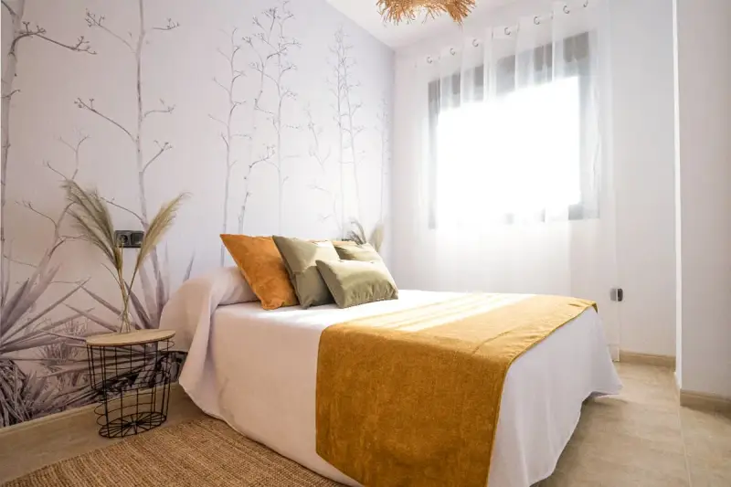 Habitación con papel tapiz de ramas y gotele moderno. Cama con cojines verdes y naranjas, y manta mostaza.