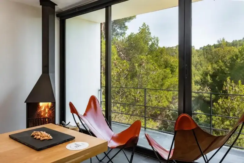 Chimenea encendida y sillones de cuero frente a un balcón con vistas a un bosque frondoso, ideal para el turismo interior España.