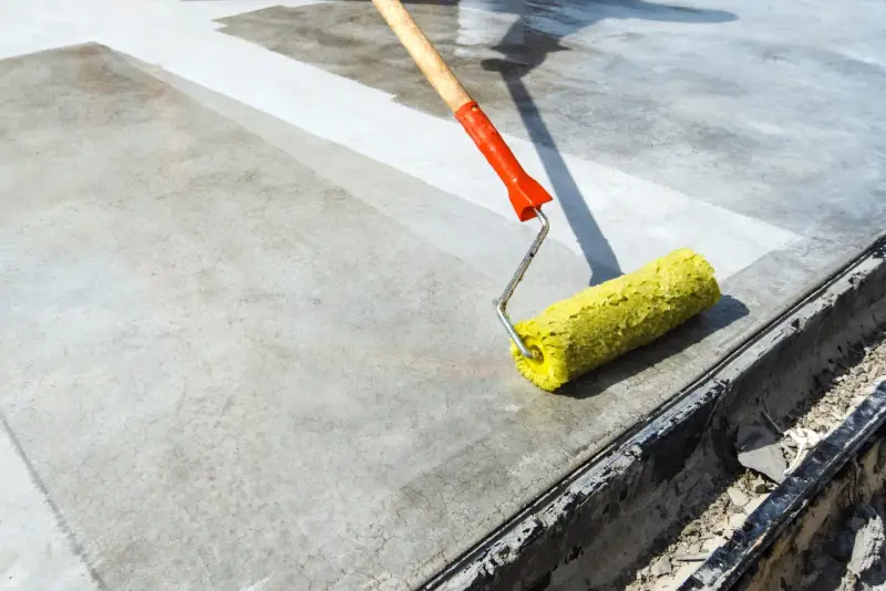 Wałek z żółtym włosiem rozprowadza środek do impregnacji betonu na szarej powierzchni.