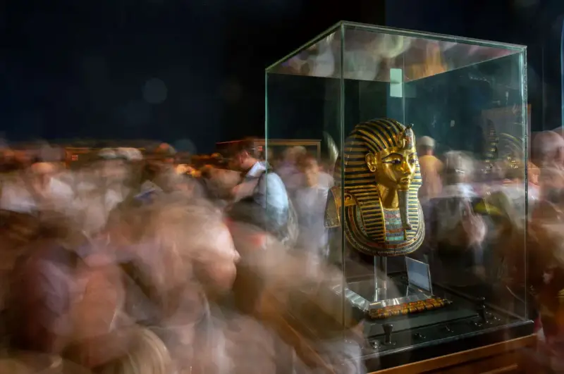 Goldene Maske des Pharao Tutanchamun in einer Vitrine, umgeben von verschwommenen Besuchern.