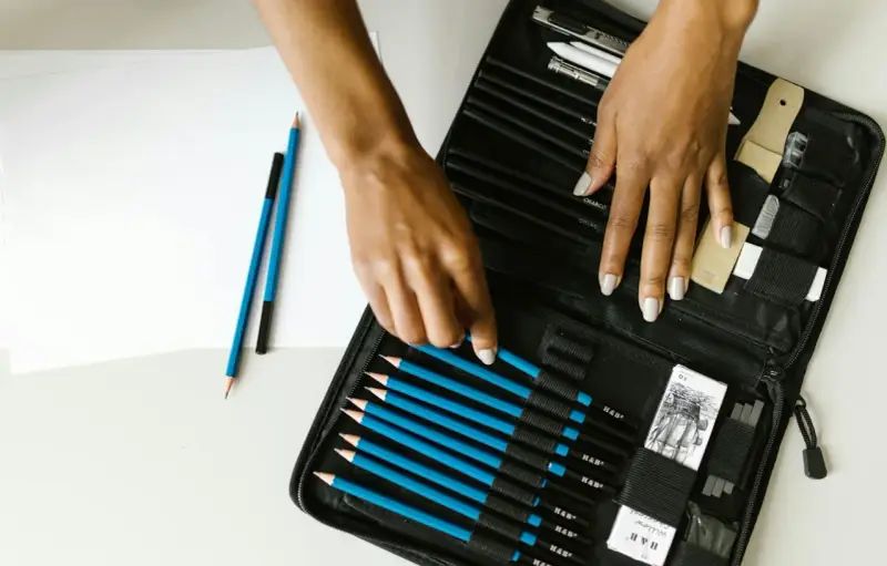 Un étui ouvert révèle une sélection de crayons de différents types pour dessiner, avec des mains choisissant un crayon bleu.