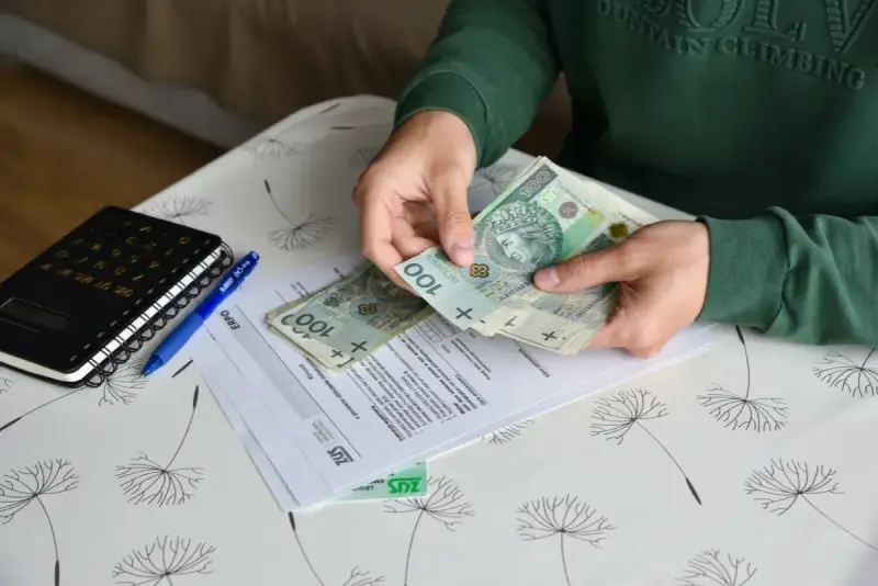 Ręce liczą banknoty 100 zł nad dokumentami ZUS. Obok leży kalkulator i długopis. To może być pomocne przy ustalaniu, ile wynosi świadczenie przedemerytalne na rękę.