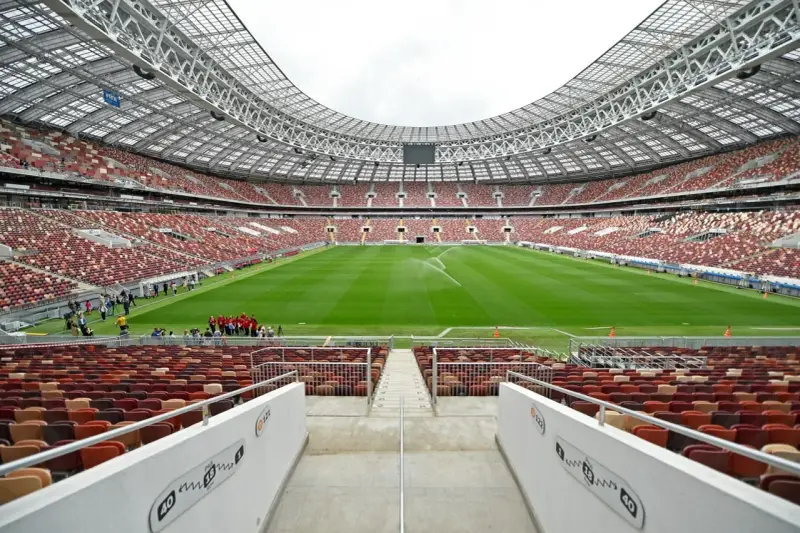 Pusta trybuna moskiewskiego stadionu z rzędem czerwonych i brązowych siedzeń. Na boisku pracują zraszacze.