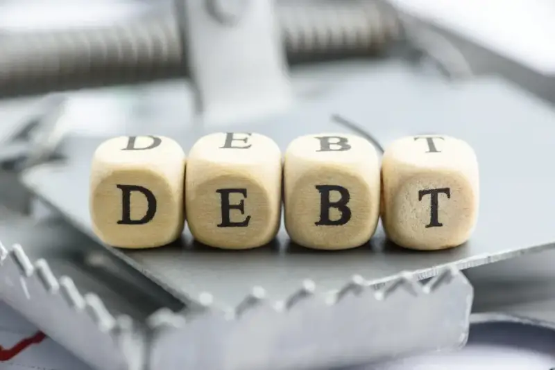 Cztery drewniane kostki z literami D, E, B, T tworzą słowo "DEBT" na pułapce. To symbol finansowych zobowiązań, które mogą prowadzić do trudnej sytuacji, jak datio in solutum.