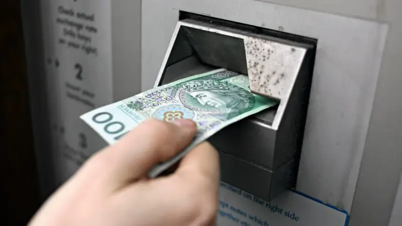 Ręka wkłada banknot 100 zł do bankomatu. Czy państwo może zabrać pieniądze z konta w razie wojny?