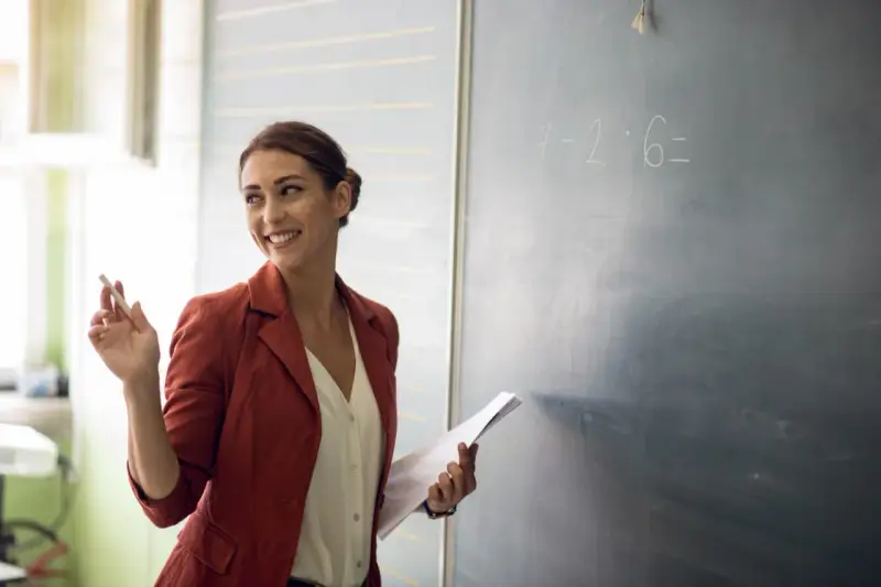 Lehrerin mit Kreide und Unterlagen vor einer Tafel mit einer Matheaufgabe. Sie lächelt und wirkt engagiert, was auf einen interaktiven Unterricht hindeutet.
