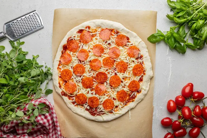 Surowa pizza z sosem, serem i okrągłymi plasterkami peperoni, gotowa do pieczenia. Obok świeża bazylia i pomidorki.