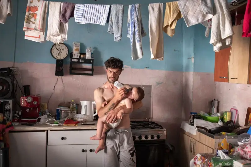 Emiliano Dante, padre single, nutre il suo bambino in una cucina disordinata, con panni stesi ad asciugare.