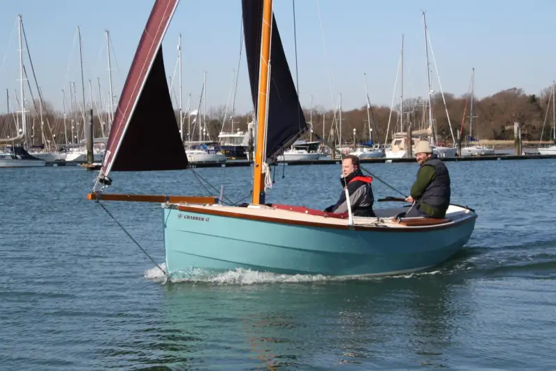 Deux hommes naviguent sur un petit bateau à voile bleu clair, avec d'autres voiliers en arrière-plan.
