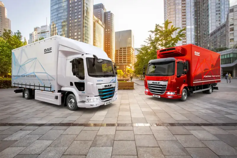 Dos camiones DAF, uno blanco eléctrico y otro rojo, en una ciudad moderna. El DAF XB blanco muestra "POWERING YOUR SUCCESS".
