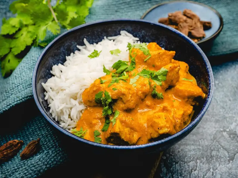 Ein cremiges Chicken Korma mit Basmatireis, garniert mit frischem Koriander. Dazu Gewürze und ein kleiner Teller mit Fleischstücken.