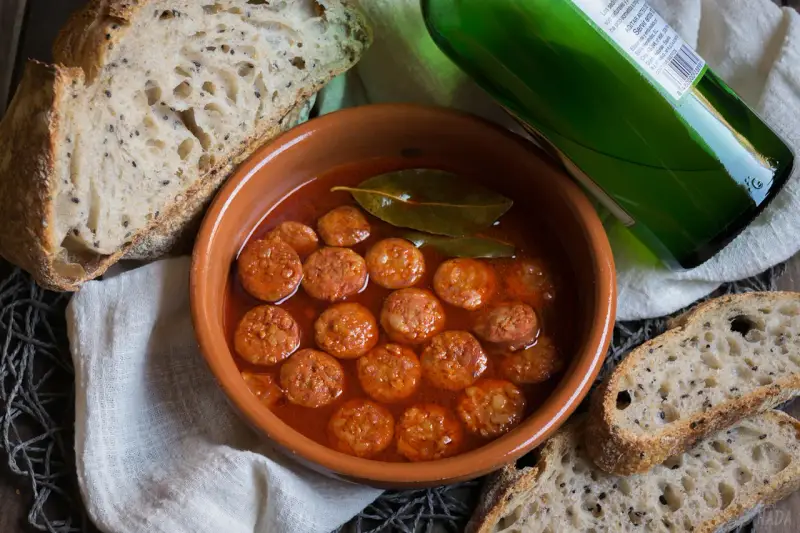 Chorizo a la sidra en cazuela de barro, acompañado de pan rústico y una botella de sidra verde.