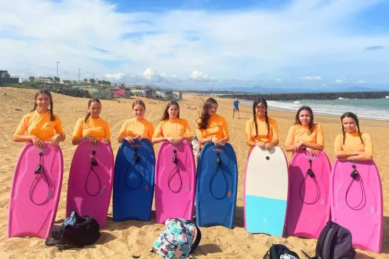 Huit jeunes filles en t-shirts orange, prêtes pour leur colonie à la mer, posent avec leurs bodyboards sur la plage.
