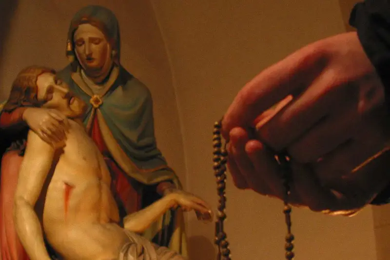 Eine Hand hält einen Rosenkranz, während die Pietà-Statue im Hintergrund zu sehen ist. Ein evangelischer Rosenkranz wird hier in Andacht gehalten.