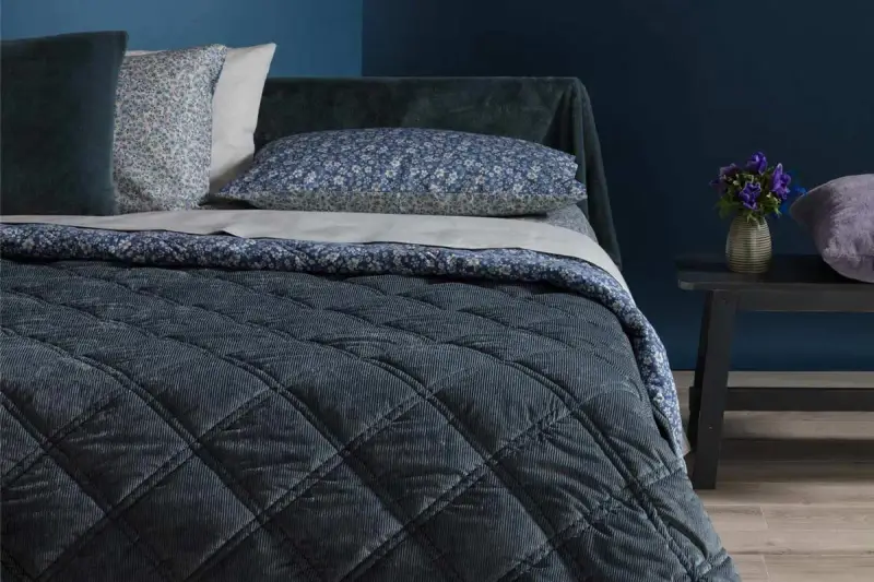 Letto con copriletto trapuntato blu scuro, cuscini floreali e un dettaglio che ricorda che il velluto si rovina con acqua.