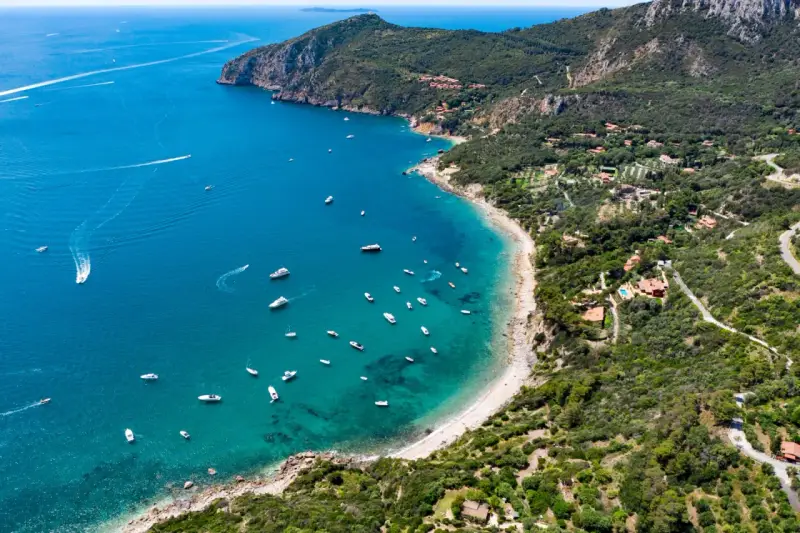Baia con acque cristalline e numerose barche, incastonata tra colline verdi e scogliere. Un vero gioiello tra le spiagge più belle Toscana.