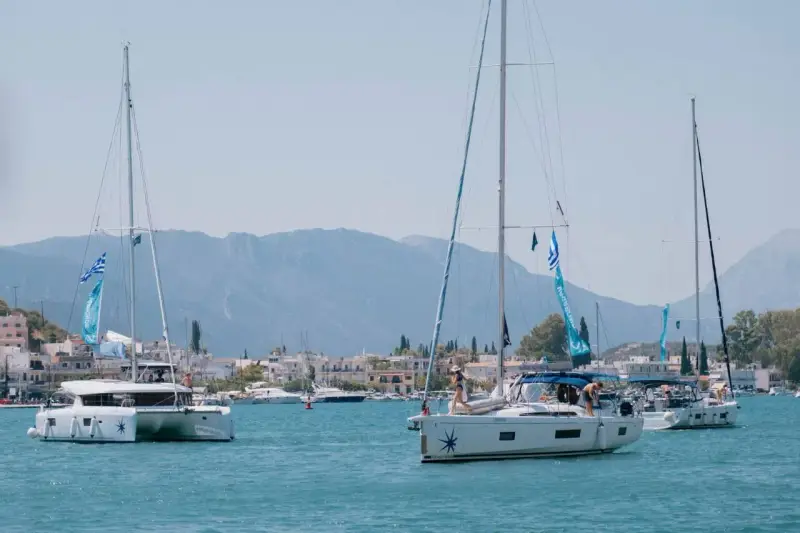 Trois voiliers naviguent dans une baie ensoleillée. Au premier plan, un catamaran monocoque blanc avec des voiles bleues et blanches. Derrière, un autre voilier blanc avec des personnes à bord. Au loin, une ville côtière et des montagnes.