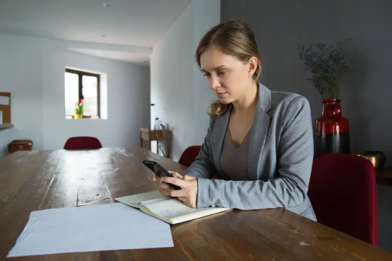 Kobieta z telefonem w ręku przy drewnianym stole. Analizuje dane, szukając odpowiedzi, dlaczego najem średnioterminowy staje się bezpieczniejszą alternatywą dla właścicieli.