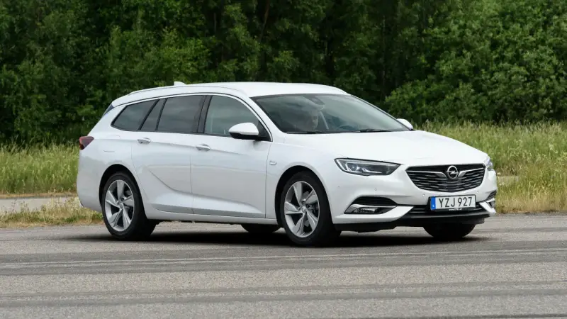 Vit Opel Insignia kombi parkerad vid en grönskande skog. En förare syns i bilen, kanske funderar på ett opel insignia problem.