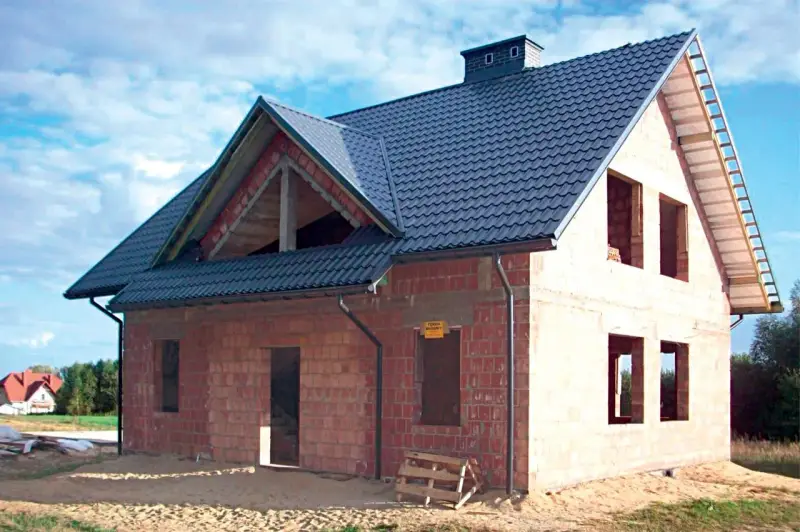 Budowa domu z cegły, widoczny dach betonowy skośny ocieplenie. Widać otwory okienne i drzwiowe, komin.