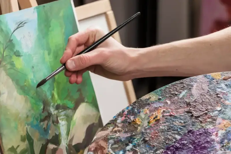 Main d'artiste peignant une scène verdoyante avec une peinture à l'acrylique. Palette de couleurs vibrantes.