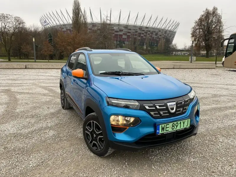 Niebieski Dacia Spring z pomarańczowymi akcentami na tle Stadionu Narodowego. Sprawdź atrakcyjną dacia spring cena!