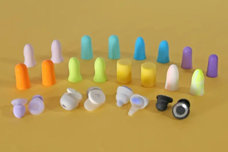 Apprenez comment mettre des bouchons d'oreilles en mousse. Divers bouchons colorés et écouteurs sans fil sont disposés sur fond jaune.