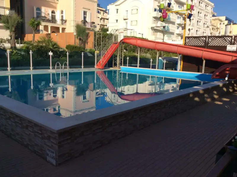 Piscina con scivolo rosso e giochi d'acqua ai Bagni Carmen Cattolica. Riflessi degli edifici nell'acqua azzurra.