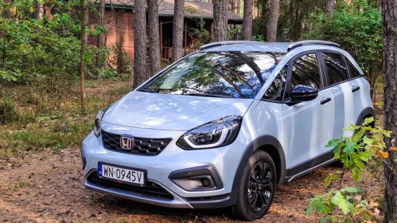 Jasny Honda Jazz Crosstar zaparkowany w lesie, obok drewnianego domku.