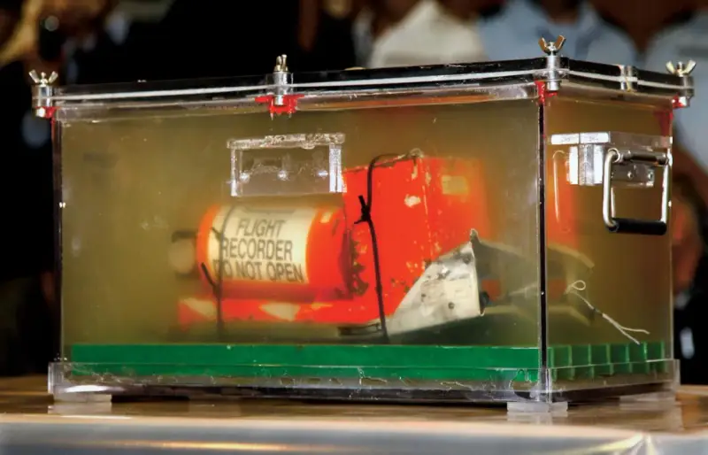 Pomarańczowa skrzynka "czarnej skrzynki" z napisem "FLIGHT RECORDER DO NOT OPEN" w przezroczystej obudowie, dowód w sprawie wypadku lotniczego.