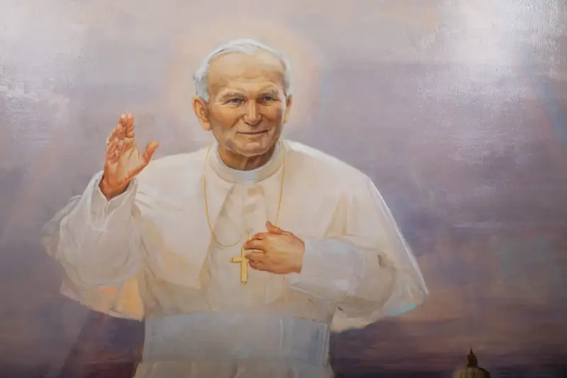 Obraz przedstawia Jana Pawła II w białej szacie, z podniesioną prawą dłonią i krzyżem na piersi. Wierni odmawiają modlitwę do Jana Pawła II o uzdrowienie.