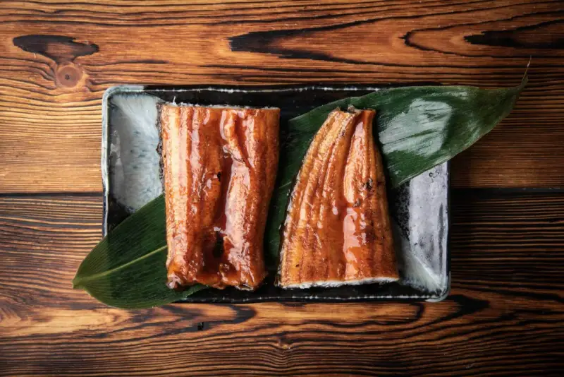 Dwa kawałki błyszczącego, glazurowanego sushi z węgorzem na liściu, podane na czarnej tacce.