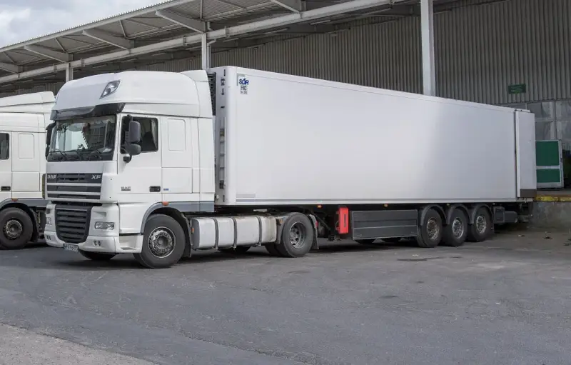 Biała naczepa DAF XF z chłodnią, gotowa do transportu. Obok stoi kolejna ciężarówka, tworząc zgrany zespół pojazdów.