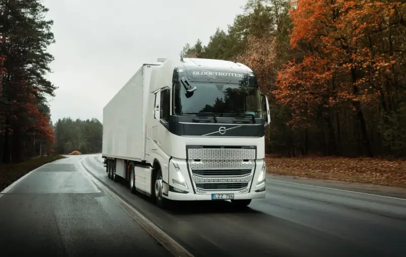 Biała ciężarówka Volvo FH12 z naczepą jedzie po mokrej drodze wśród jesiennego lasu.