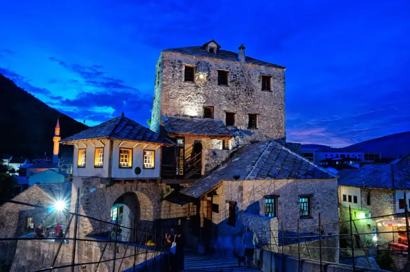 Kamienne budynki i most w Mostarze o zmierzchu, z widocznym minaretem w tle.