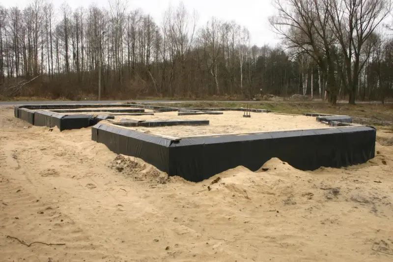 Gotowe ławy fundamentowe otoczone czarną folią, czekają na zalanie betonem. W tle las.
