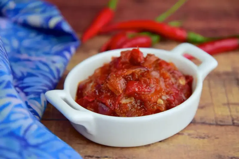 Pikantny sos sambal oelek w białej miseczce, obok czerwonych papryczek chili i niebieskiej tkaniny.