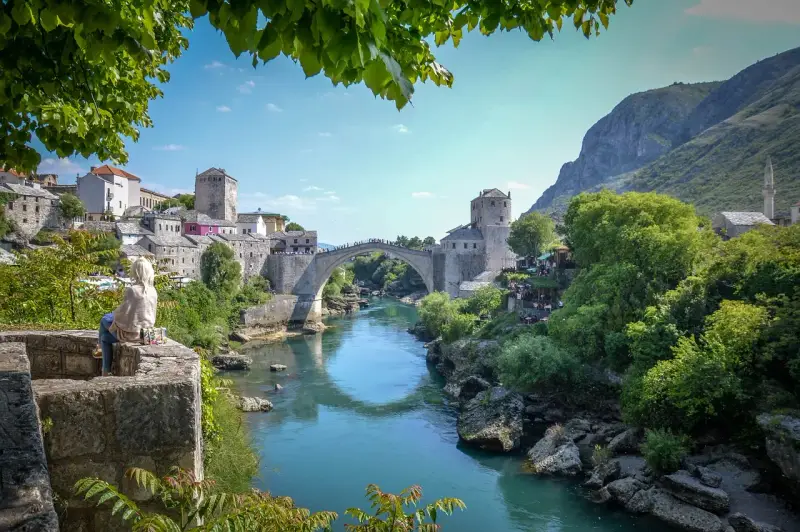 Widok na Mostar w Bośni z kamiennym mostem Stary Most nad turkusową rzeką Neretwą. W tle kamienne budynki i góry.