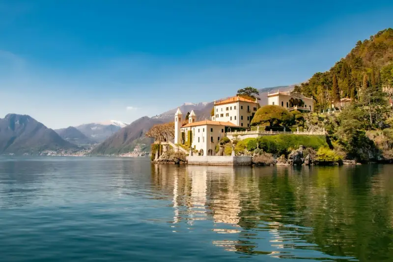 A scenic journey from Milan to Lake Como reveals this stunning villa on the water's edge, with mountains in the distance.
