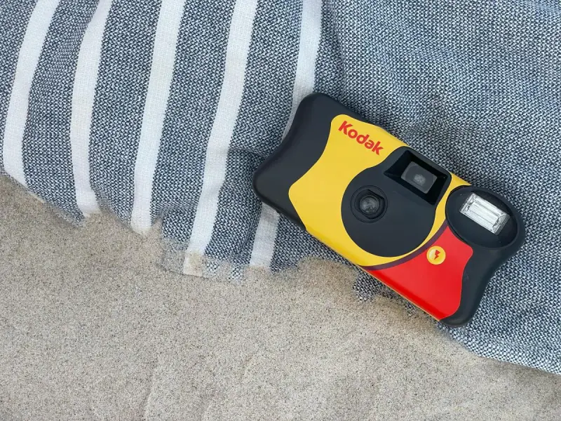 Aparat jednorazowy firmy Kodak leży na plaży obok ręcznika. Idealny do uchwycenia letnich wspomnień.